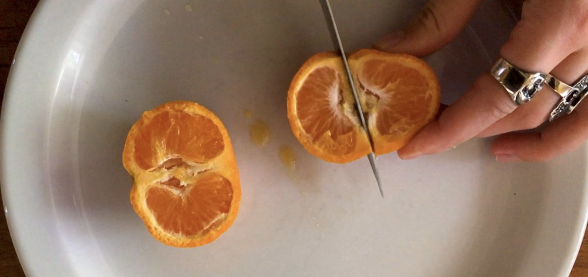 A hand with ringed fingers holds half a mandarine orange that is being sliced on a white plate