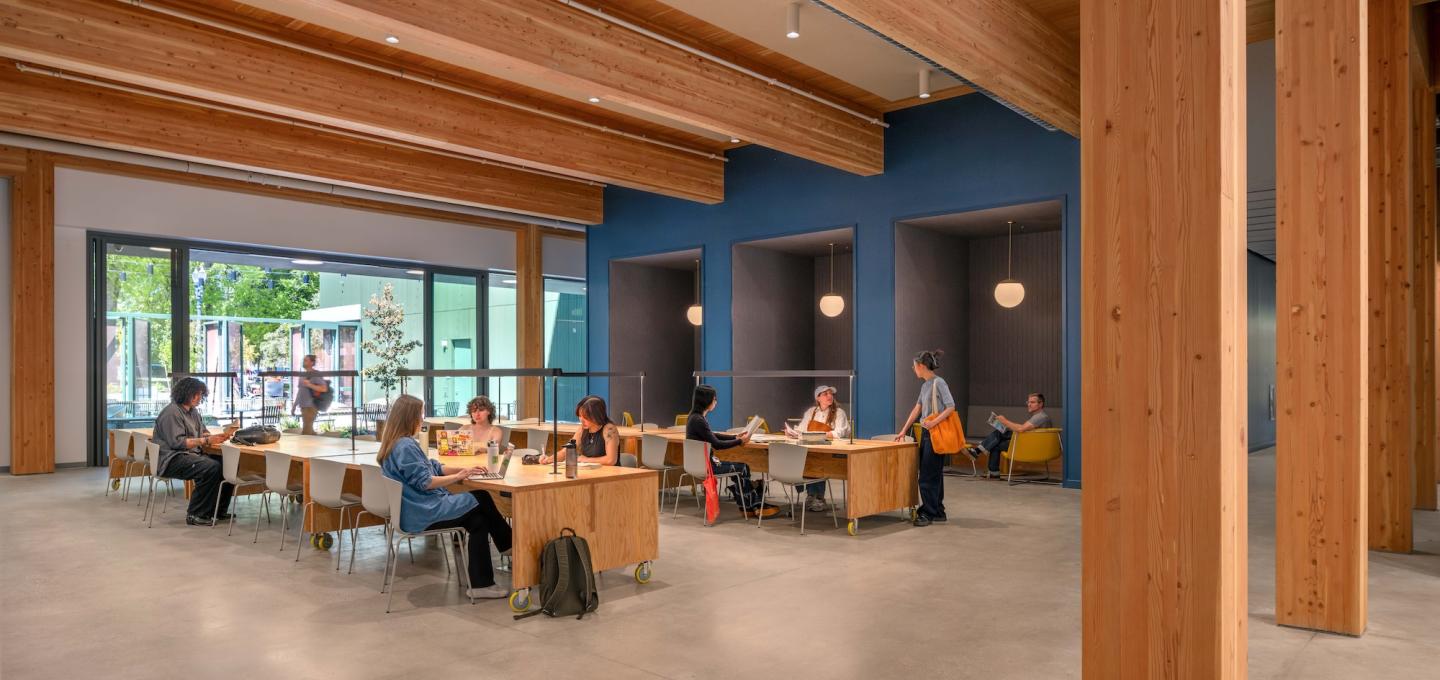 Students gather at tables in a large open room with wooden beams and columns and three recessed study nooks. A large sliding glass room is open to an gated outdoor work yard.