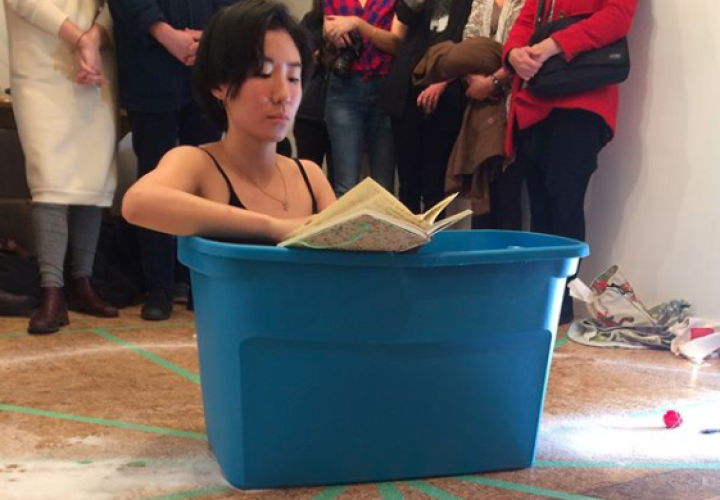 Woman seated in blue plastic tub while reading poetry, performance at an art show reception.