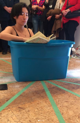 Woman seated in blue plastic tub while reading poetry, performance at an art show reception.