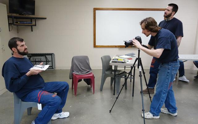 Man with camera on tripod films a seated man while another looks on.