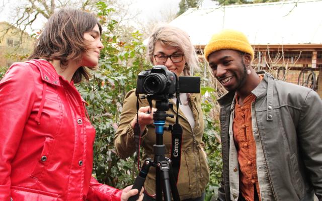 Three students in a video and community course gather around a camera on a tripod