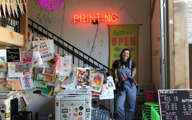 Kate Bingaman Burt stands next to a Risograph printer under a neon sign that says "Printing"