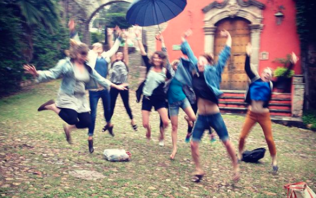 Group of students, one holding an umbrella, jump exuberantly into the air. They are assembled in a brick and moss covered courtyard in front of a red building in Guanajuato, Mexico