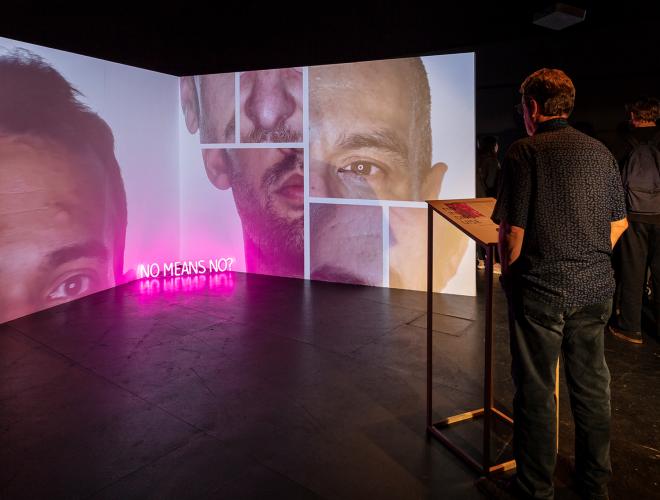 Man stands at a podium in front of a video projection with collage of male faces