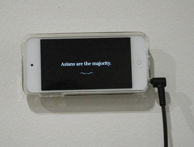 iPhone mounted on wall with headphones attached for interactive experience. The screen says "Asians are the majority."