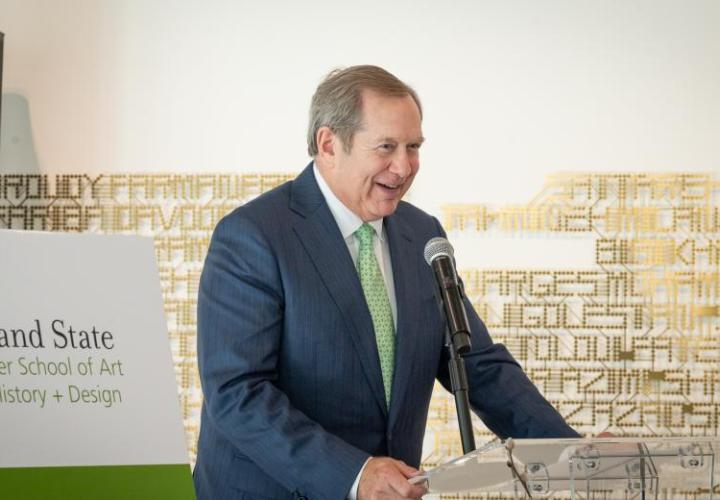 A man in a suit stands at a podium in front of a microphone, smiling