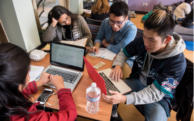 Image of students gathered around a table on their phones and laptops. 