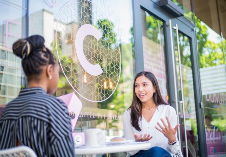 two people talking at a table outside in front of a large glass window