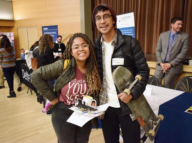 Two students dress casually at PSU Career Fair. One student is holding a handfull of free goodies and the other is holding a skateboard.