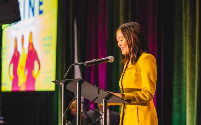 woman in a yellow blazer speaking at a podium