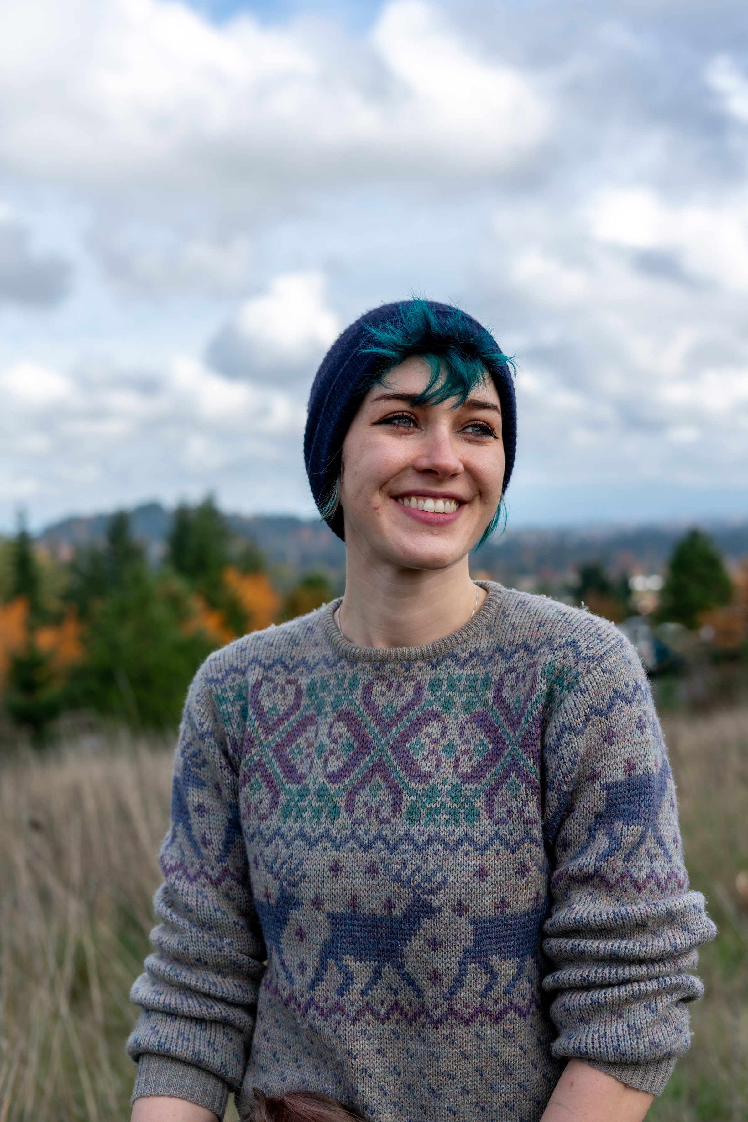 Professional photo of Sloan Oakrest, a white person with dyed teal hair. They are wearing a black beanie and a grey sweater with intricate knit patterns. They are looking off to the right with a wide, open mouthed smile in front of a blurred backdrop of grass, trees and a blue sky with clouds. 