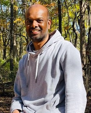 A photo of Sivanna Chithanna, a dark-skinned person with short grey hair and beard. They are wearing a light grey hoodie and standing in front of some trees with yellow and green leaves. 