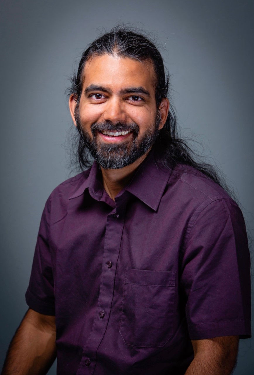Sachin Nedungadi, a brown skinned man with long black hair, a beard and a purple shirt, smiling at the camera.