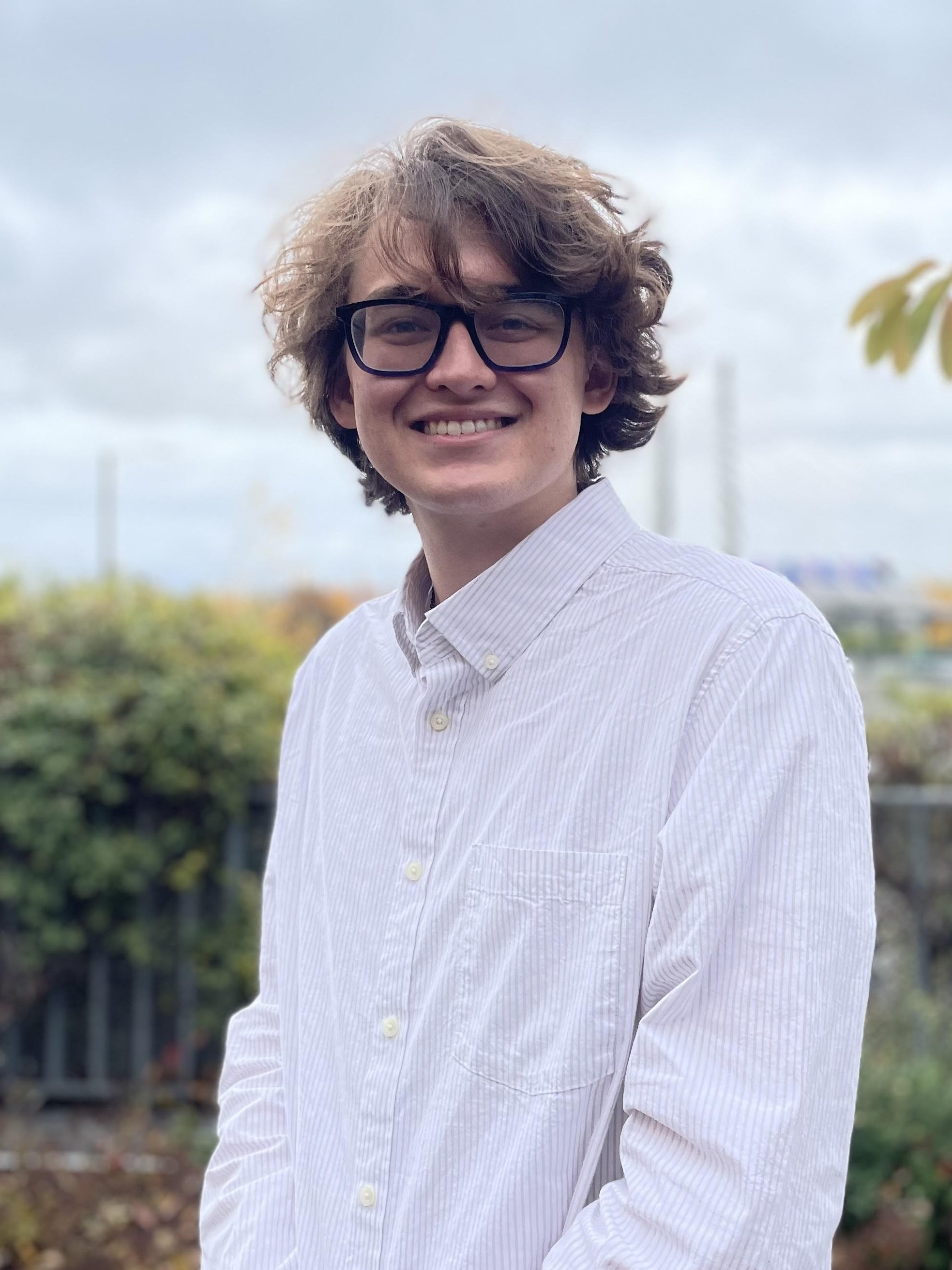 A professional headshot of Tim McWilliams, a young white man with midlength wavy brown hair and glasses.