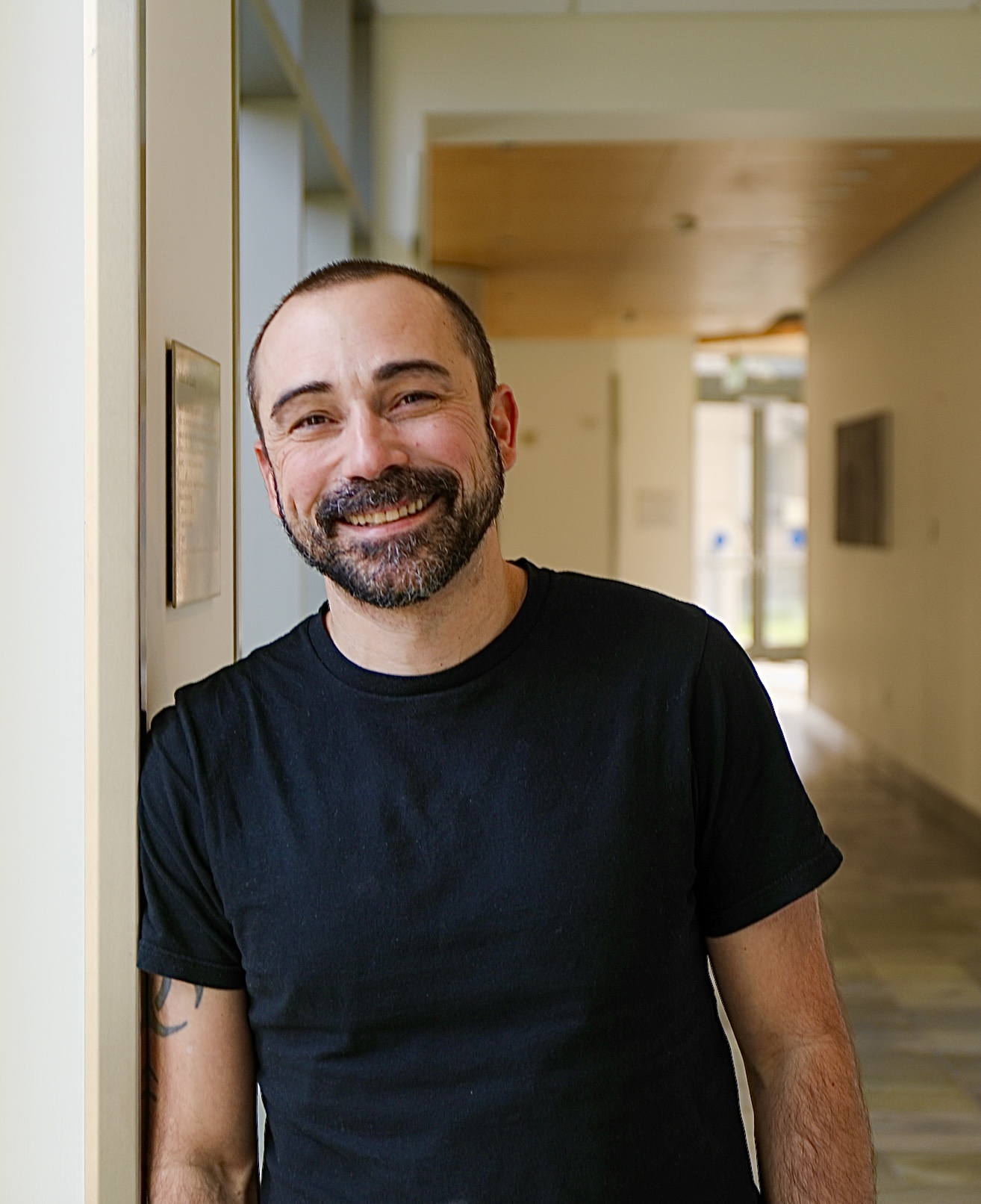 Michael Cohen, a white person with shorn black hair and a beard. They are leaning against a doorframe and smiling at the camera.