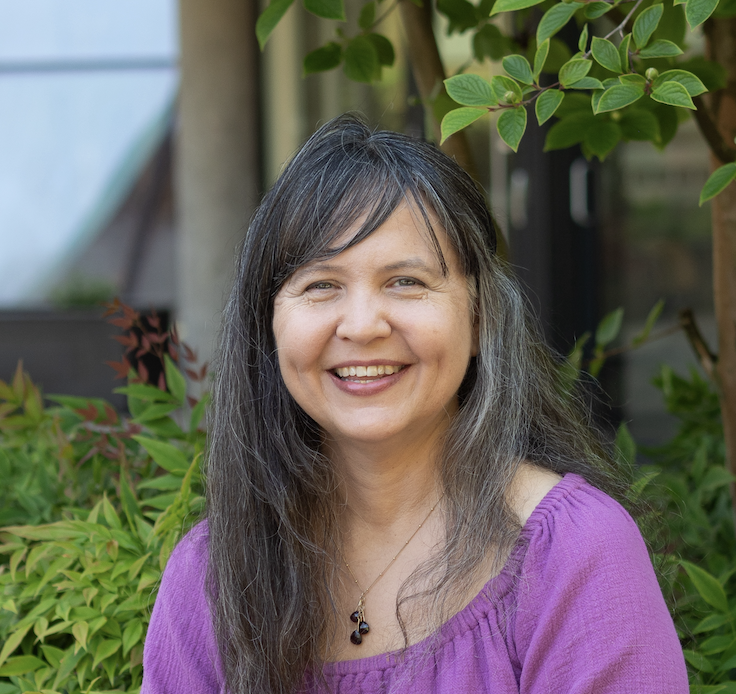 A professional headshot of Ashlie Sarsgard, a white woman with long black and grey hair sitting outside amongst some greenery. 