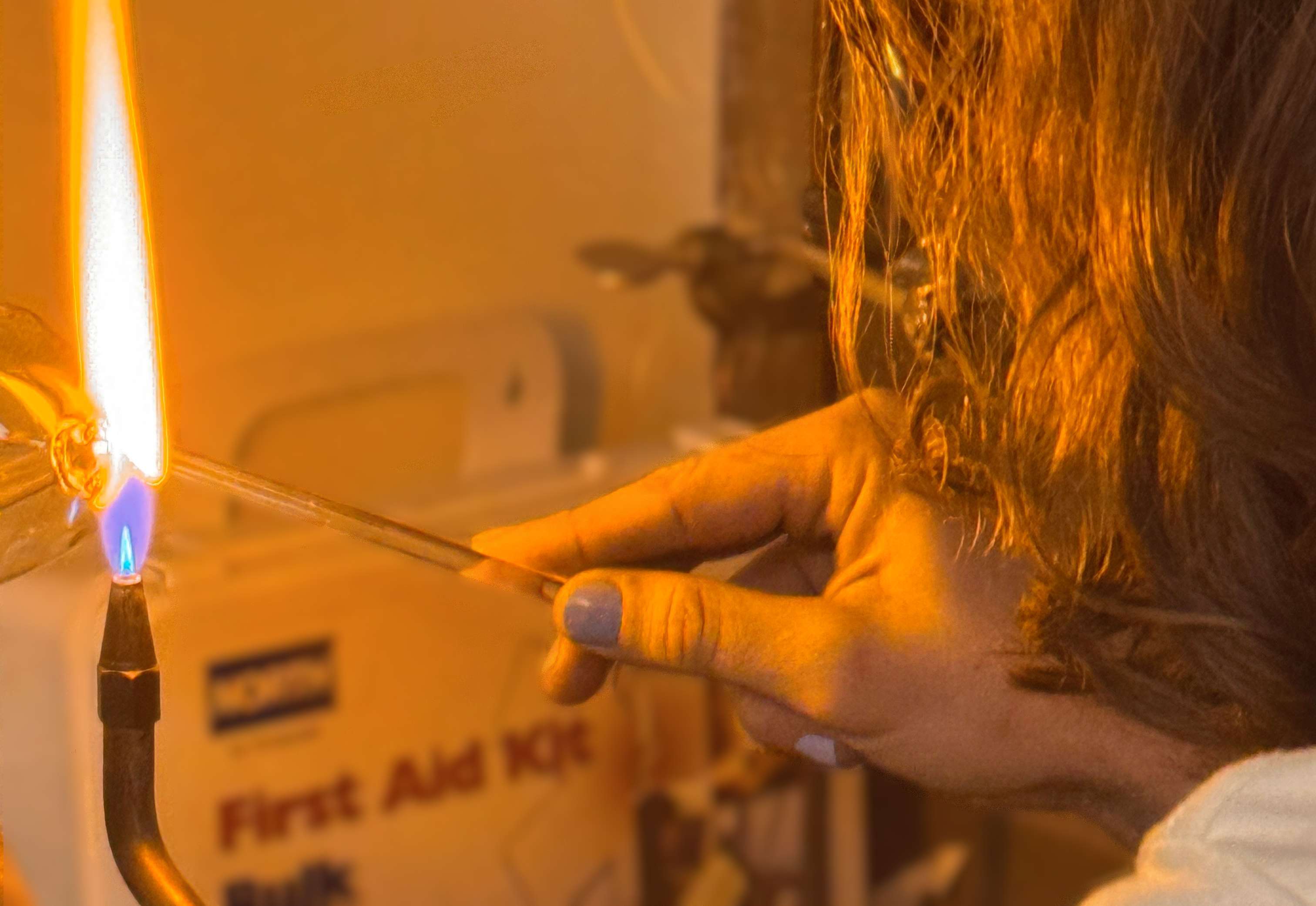Image of white student with blue nail polish and midlength hair lifting a glass rod to a glass blowing flame tool. 
