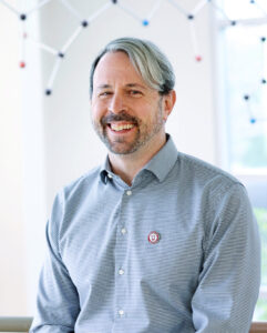 Matthew Sigman, a white man with short wavy grey hair. There are chemical models scattered in the background behind him.