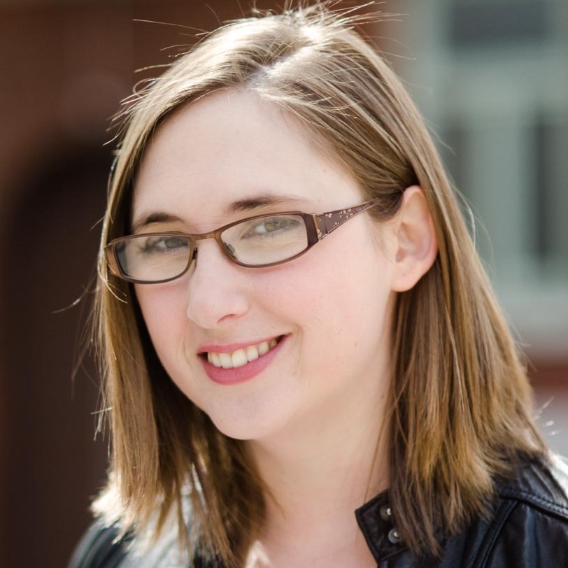 A professional portrait of Theresa McCormick, a white woman with mid-length brown hair, wearing a black leather jacket and brown, rectangular framed glasses.