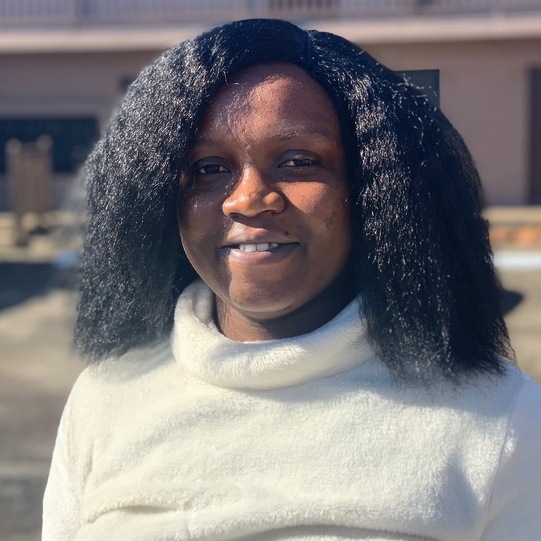 Ewubarena, a black woman with mid-length black hair and brown eyes, is wearing a white fluffy sweater and smiling. 