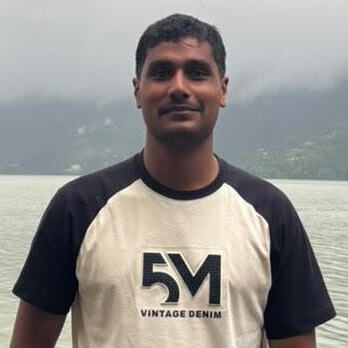 Nabin Kumar Thapa, a tan-skinned man wearing a white t-shirt with black sleeves and text at its center. He has short black hair and is standing in front of a body of water with fog cover. 