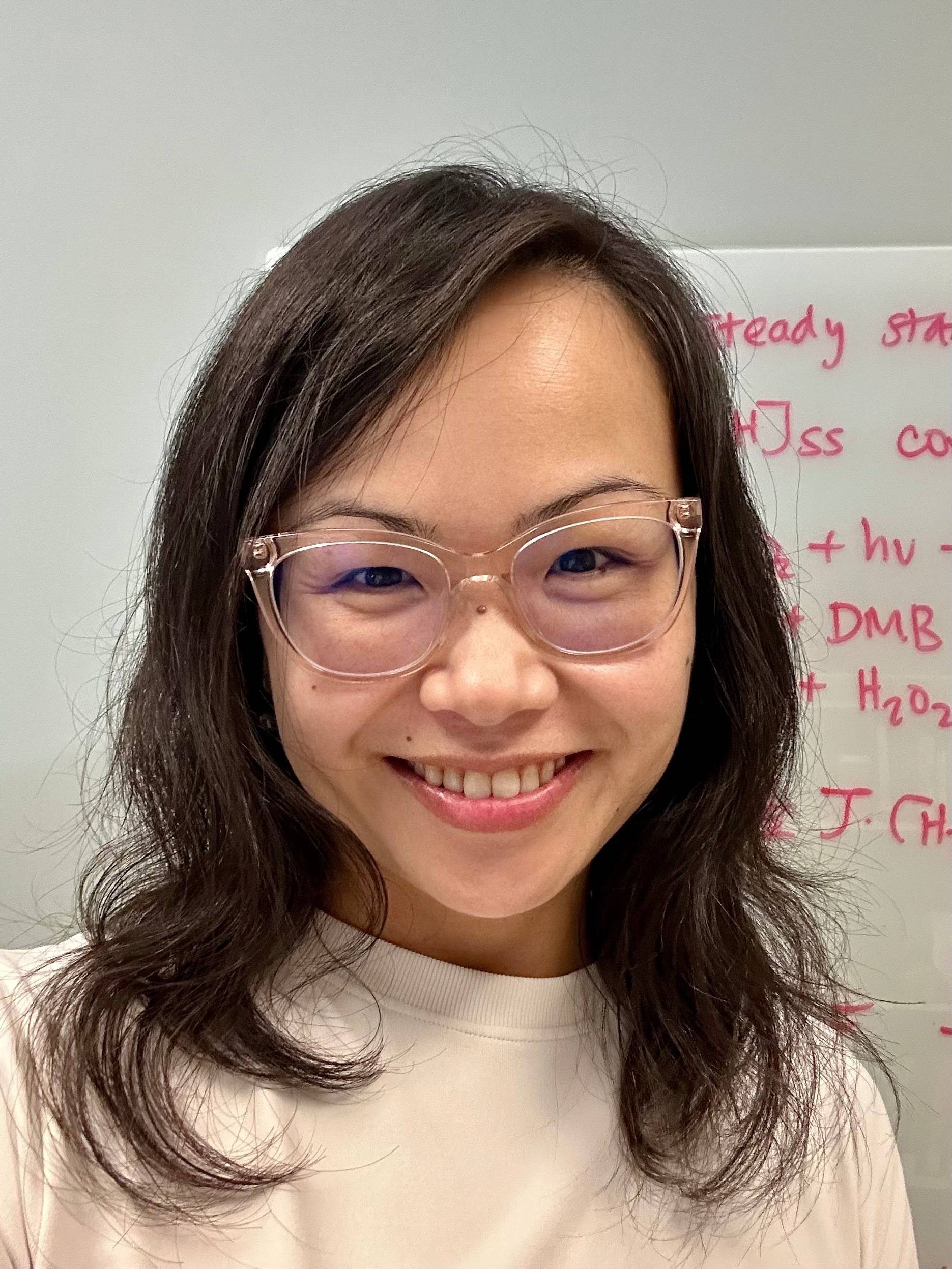 A selfie portrait of an asian woman wearing clear framed glasses and medium length hair. She is smiling and standing in front of a whiteboard with red text on it.