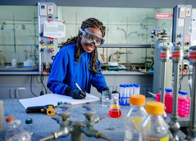 Student working in lab smiles at camera while writing in notebook. 