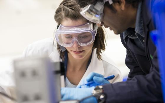 A TA demonstrates something off camera to a student; both are in lab PPE.