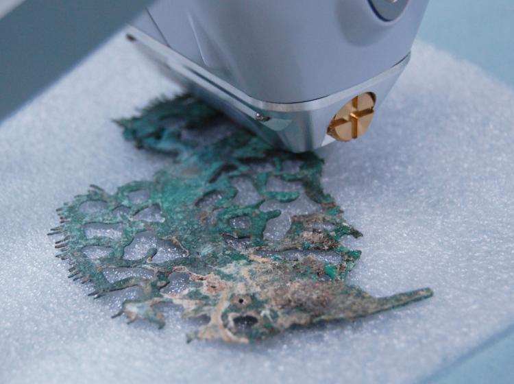 A closeup of a flat, intricate copper object covered with green corrosion being scanned by a handheld instrument