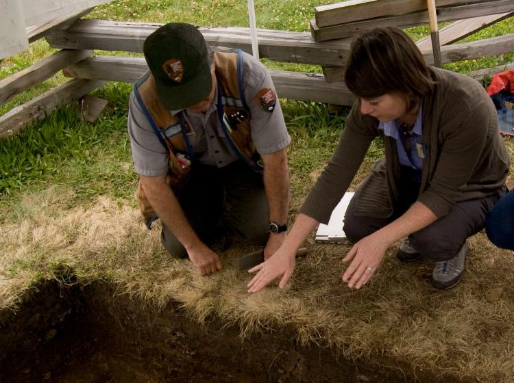 Two figures squatting on grass near a square dirt hole