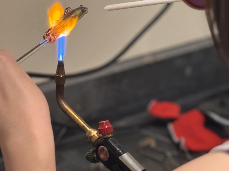 A photo of a white student using a white stick to adjust a glass piece over an blue torch flame. 