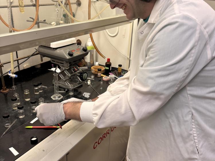 Kieran, a white student with midlength hair, is smiling and working on some glass pieces under a fume hood. 