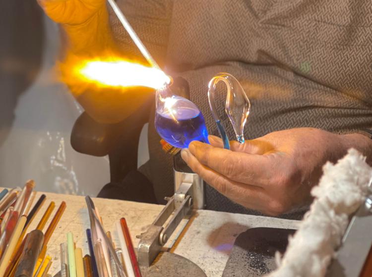 Student is using a blowtorch on a teardrop shaped glass piece with both clear and deep blue glass. 