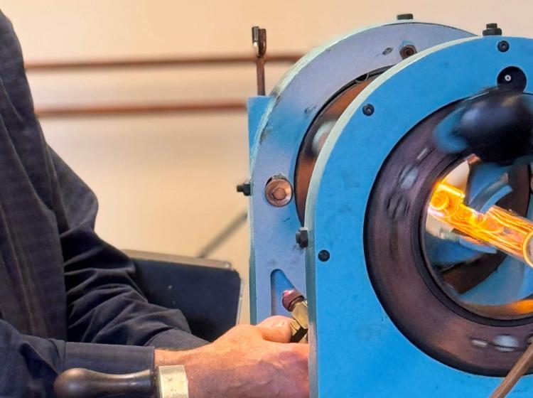 Photo of a white elder student using a glassblowing machine that is a horizontal tube-type structure with a bright blue coloration for the main body, a black internal ring and then an internal glass that is filled with orange flame.