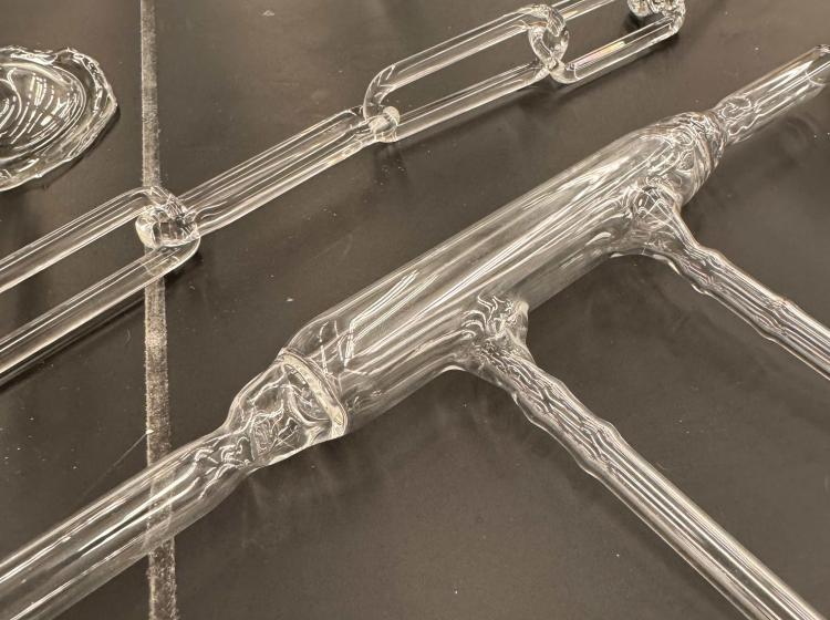 Diagonal overhead view of black tabletop with glassware pieces laid out on it. The primary two in frame are a glass chain link and then a glass tube with two attached legs perpendicular to the wide base tube. 