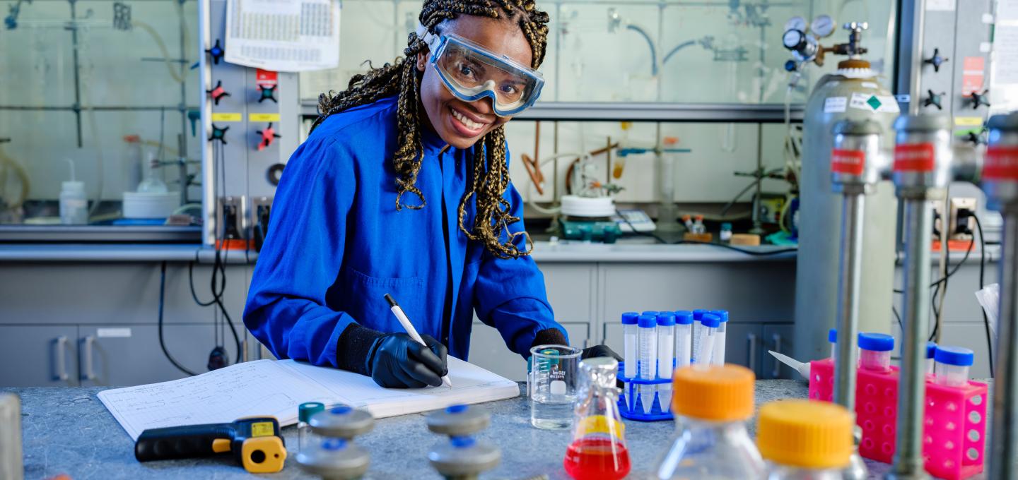 Student working in lab smiles at camera while writing in notebook. 