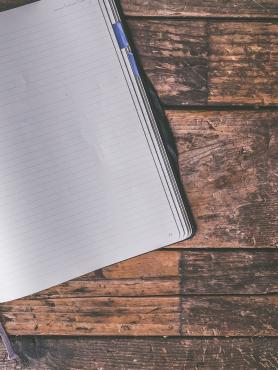 A pen sits in a lined notebook which is laid open on a wooden table. 