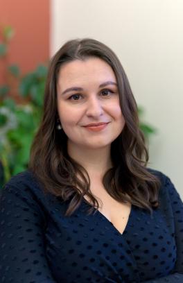 M. Popova headshot, a woman with long wavy brown hair. She is wearing a v-neck navy blouse. She is facing the camera, turned slightly toward the right, and she is smiling close-mouthed in front of a blurred green, red and off-white background..