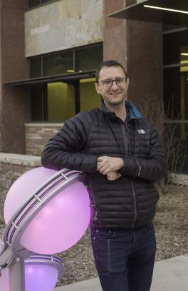 Andy McNally, a white man with short brown hair and glasses, looking toward the camera with his arms crossed. He is leaning against a glowing purple sphere in a black puffer jacket.