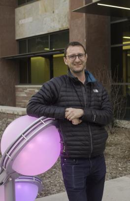 Andy McNally, a white man with short brown hair and glasses, looking toward the camera with his arms crossed. He is leaning against a glowing purple sphere in a black puffer jacket.