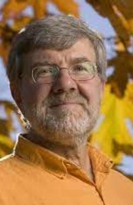 Carl Wamser headshot. Carl is a white man with grey hair and a mustache and beard. He facing the camera, slightly angled to the right, with a closed mouth. He is wearing glasses and an orange buttondown shirt. Behind him is a blurred background of orange and yellow fall leaves against a light blue sky. 