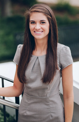Molly Atkinson, a thin white woman with long brown hair is smiling at the camera in a grey dress.