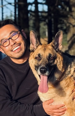 Lei Wang, a young asian man with short black hair and glasses, is seated with a german shepard dog. Both are looking at the camera. 