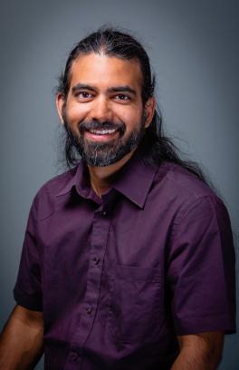 Sachin Nedungadi, a brown skinned man with long black hair, a beard and a purple shirt, smiling at the camera.