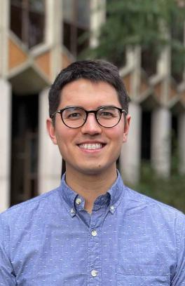 A professional portrait of Douglas Reed, a person with short black hair, light tan skin and brown eyes. He is wearing round black rim glasses and a periwinkle blue collared shirt.