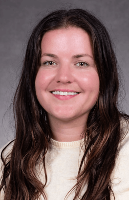 Morgan Balabanoff, a white person with long brown hair and brown eyes, smiling in a professional portrait with grey background.