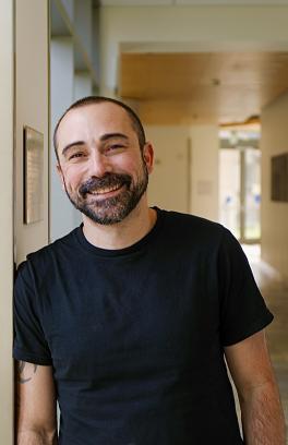 Michael Cohen, a white man with a shorn head and a full beard of black hair, is leaning his should against a wall in a black t-shirt and smiling at the camera