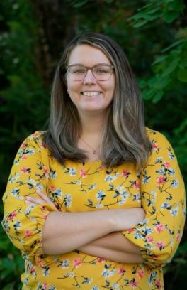 Amanda Cook, a white woman with long brown hair and glasses, is wearing a yellow blouse with flowers in front of a backdrop of greenery.