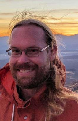 Jonathan Gross, a white man with a blonde beard and long hair, is wearing glasses and a raincoat. He's smiling in front of a scenic view of hills and a sunset.